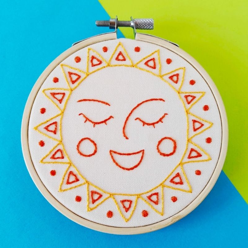 Visage de soleil souriant brodé à la main avec des fils jaunes et orange sur du lin blanc, encadré dans un cerceau de bambou